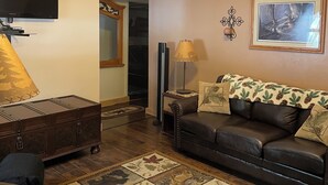 A cozy living room with a wooden ceiling, a ceiling fan, a fireplace, a leather sofa, a television, and a patterned rug. - Cozy Creekside Cottage  (Custer)