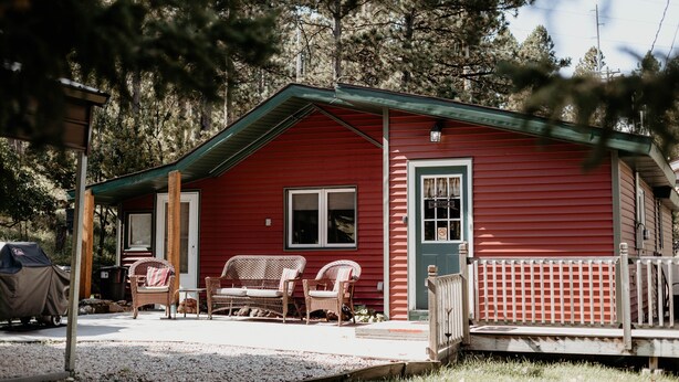 A red cabin with a porch, two wicker chairs, and a grill. - Cozy Creekside Cottage  (Custer)