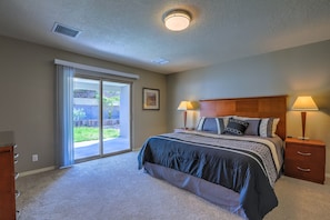 A bedroom with a large bed, two bedside lamps, a wooden headboard, a sliding glass door to a patio, and a small wooden nightstand.