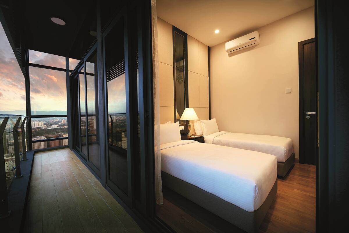 Ein Hotelzimmer mit zwei Betten, einem Nachttisch, einer Lampe und Blick auf die Stadt durch große Fenster.