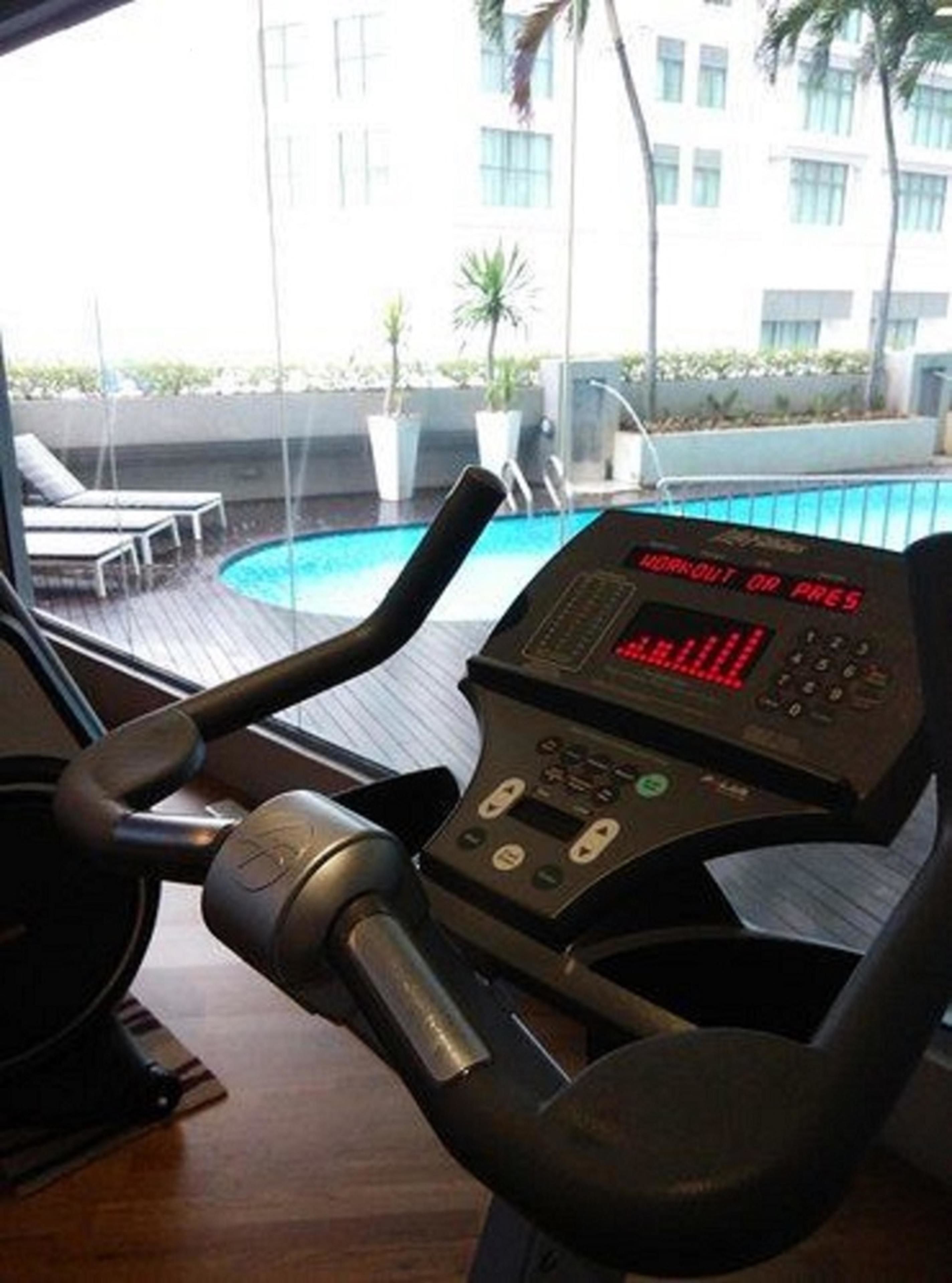 Ein stationäres Fahrrad mit digitalem Display, ein Holzboden und im Hintergrund ein Pool mit Liegestühlen.