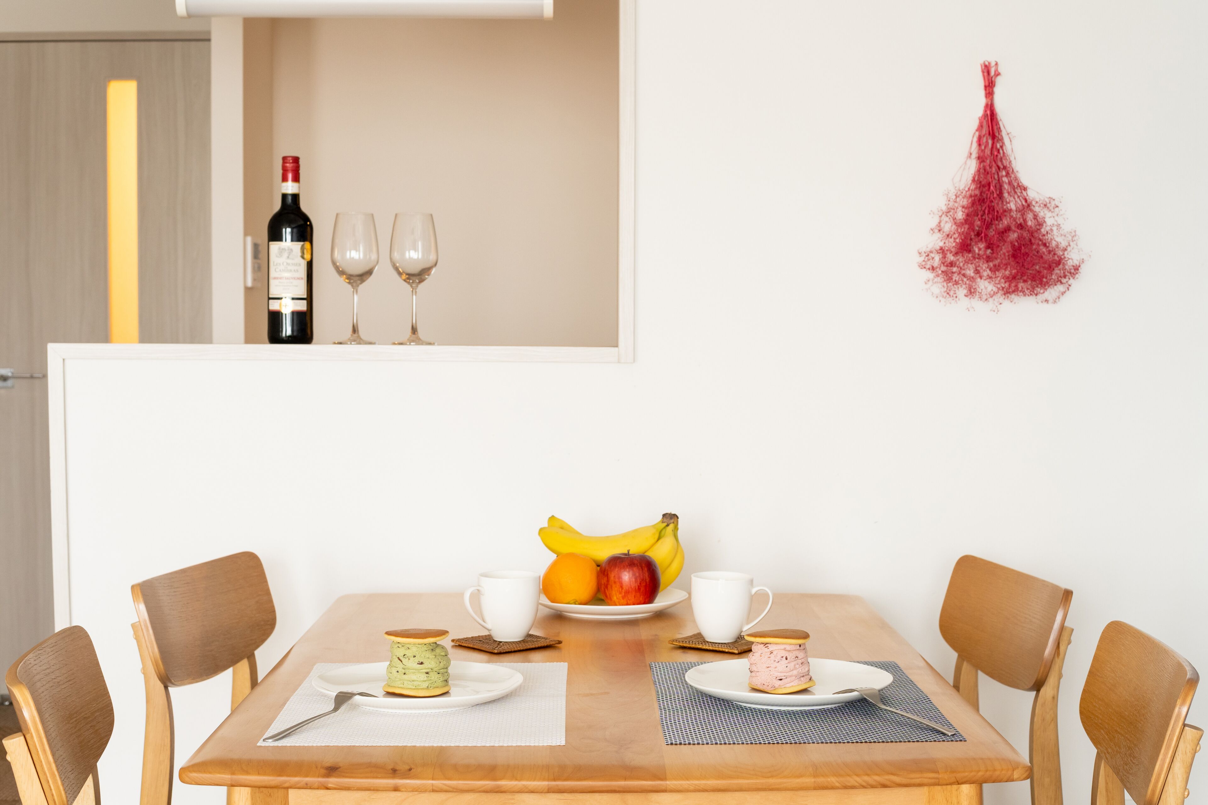 A dining table with chairs, a bottle of wine, two wine glasses, and a fruit bowl with bananas and apples.
