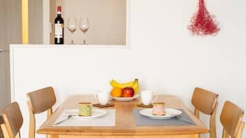A dining table with chairs, a bottle of wine, two wine glasses, and a fruit bowl with bananas and apples.