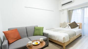 A modern hotel room with a grey sofa, a wooden bed, a small round table with a plate of fruit and two cups, and a window with curtains.