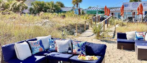 Et loungeområde ved stranden med fletningsmøbler, en køleboks med drikkevarer og udsigt til stranden og resortbygninger.