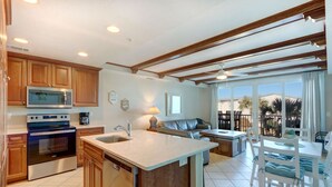A kitchen with wooden cabinets, a built-in microwave, and a dishwasher. There is a dining area with a table and chairs, and a view of palm trees through large windows. - Summer & July 4th Savings at Beachside Colony Tybee's ONLY Oceanfront Resort 2/2 (Tybee Island)