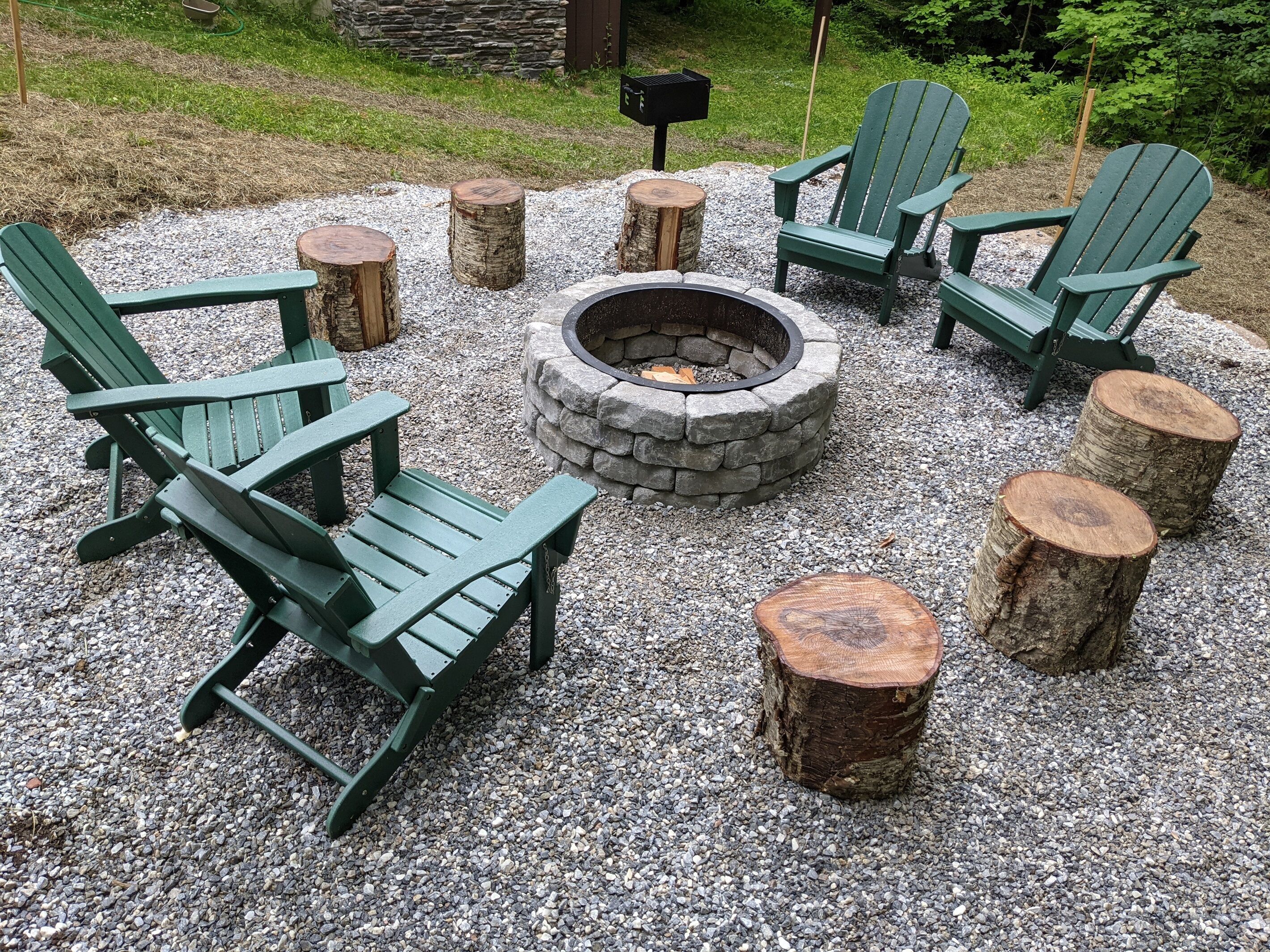 A fire pit surrounded by Adirondack chairs and wooden stools on a gravel surface.