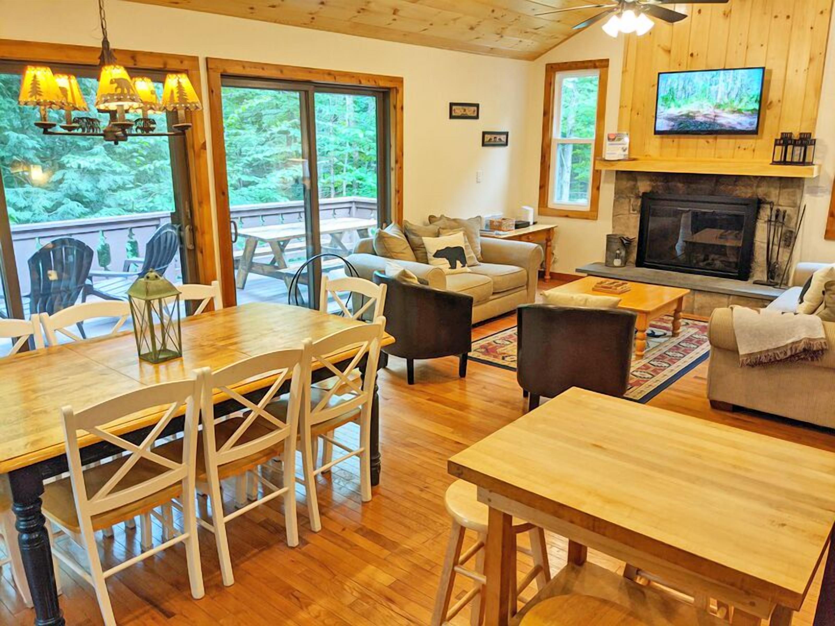 A cozy living room with a fireplace, wooden furniture, and large windows overlooking a deck.