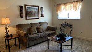 A living room with a sofa, a coffee table, a lamp, a framed picture, a window with curtains, and a small table with candles.