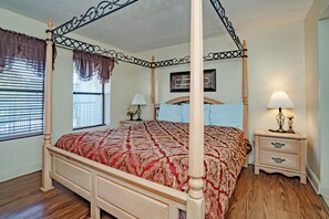 Una cama con dosel con un edredón estampado rojo, una mesita de noche con una lámpara y una ventana con persianas.