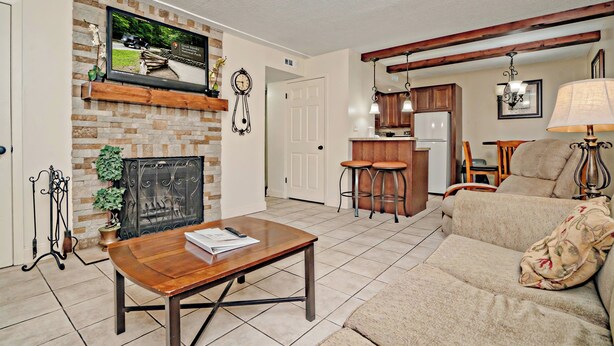 Una sala de estar con chimenea, televisor montado arriba, comedor y cocina al fondo, además de un sofá con cojín.