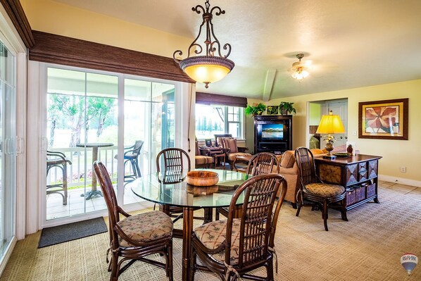 A dining area with a glass table, chairs, a chandelier, and a television in the background. - Top Floor Condo Close to Beaches (Princeville)