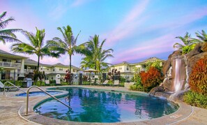 A swimming pool with a waterfall feature, surrounded by palm trees and a building with a balcony. - Top Floor Condo Close to Beaches (Princeville)
