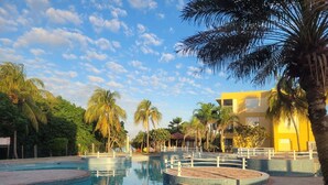 Una piscina con diverse sezioni, circondata da palme e un edificio giallo.