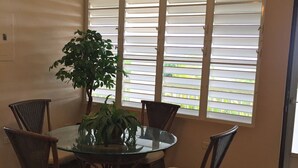 Zona pranzo con un tavolo rotondo in vetro, circondato da sedie in vimini, una ventola a soffitto e una finestra con persiane.