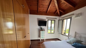 Un dormitorio con techo de madera, una cama con almohadas, un aparador, un televisor y una ventana con vistas al verdor.