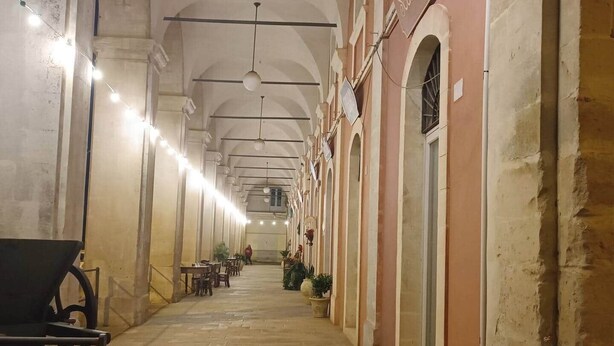 A corridor with a long bench on the left, tables and chairs along the right, and a person walking in the distance. - Old Market House Antò Ragusa (Ragusa)