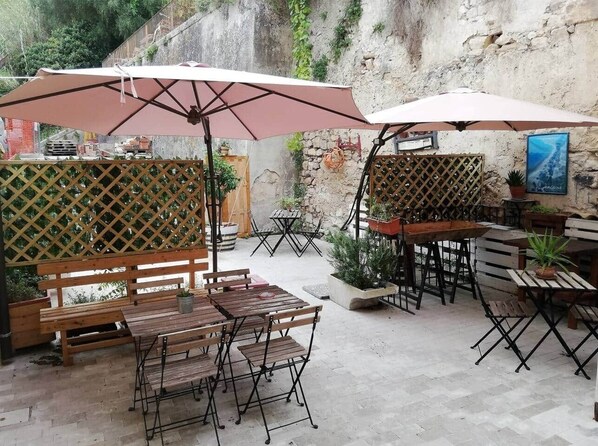 An outdoor patio area with wooden tables and benches, some with umbrellas. There are potted plants and a stone wall in the background.