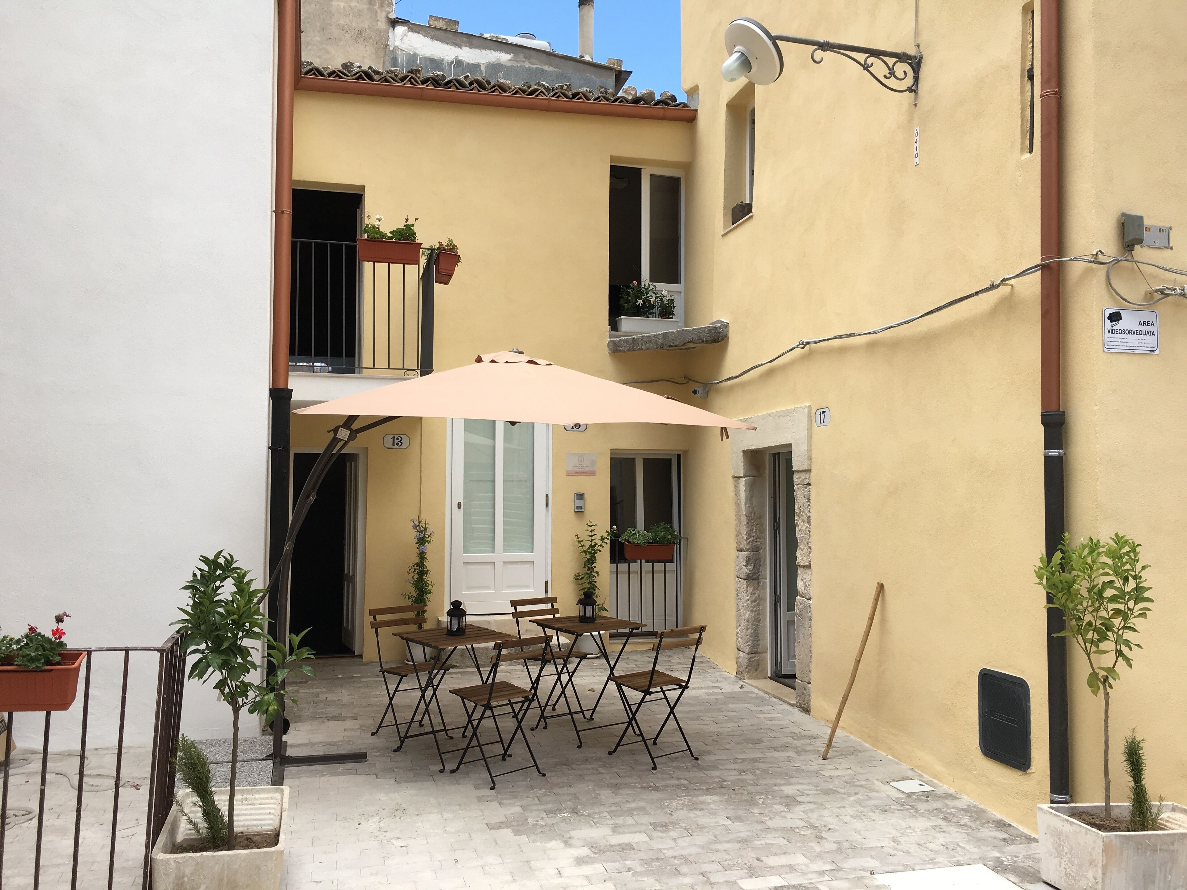 Une terrasse extérieure avec une table et des chaises, entourée de plantes en pot et d’un bâtiment jaune.