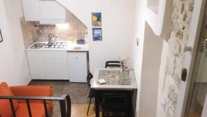 A compact kitchen with a gas stove, sink, and white cabinets. There is a dining table with chairs and a small rug. An orange sofa is visible in the foreground. - Old Market House Antò Ragusa (Ragusa)