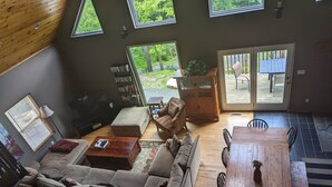 Amplia sala de estar con sofá, mesa de centro de madera y mesa de comedor con sillas. Grandes ventanas ofrecen luz natural y vistas al exterior.