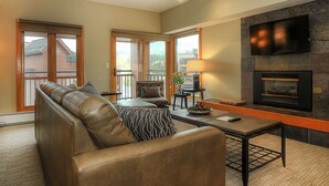 A spacious living room with a leather sofa, a fireplace, a flat-screen TV, and large windows offering a view of a building and trees. - Village Allure (Breckenridge)