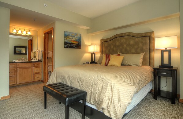 A bedroom with a large bed, two bedside tables with lamps, a bench at the foot of the bed, a dresser, a mirror, and a painting on the wall. - Village Allure (Breckenridge)