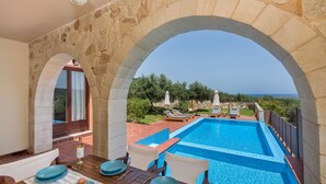 Un espace aménagé au bord de la piscine, avec une arche en pierre, une table à manger dressée avec des assiettes et une bouteille, et une vue sur la mer.