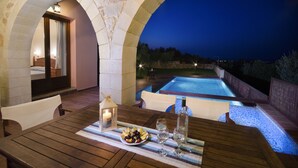 Une table en bois, ornée d’une bougie, d’une bouteille et d’une assiette de fruits, donnant sur une piscine illuminée la nuit.