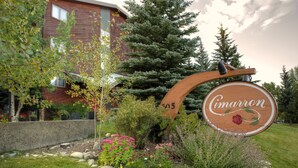 Un letrero de madera con el nombre 'Cimarron' y el número '305' frente a un edificio de pared de piedra y un jardín con diversas plantas y flores.