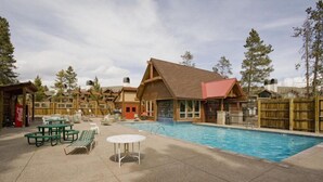 Zona de piscina con sillas de descanso y mesas, rodeada de cabañas de madera con techo rojo.