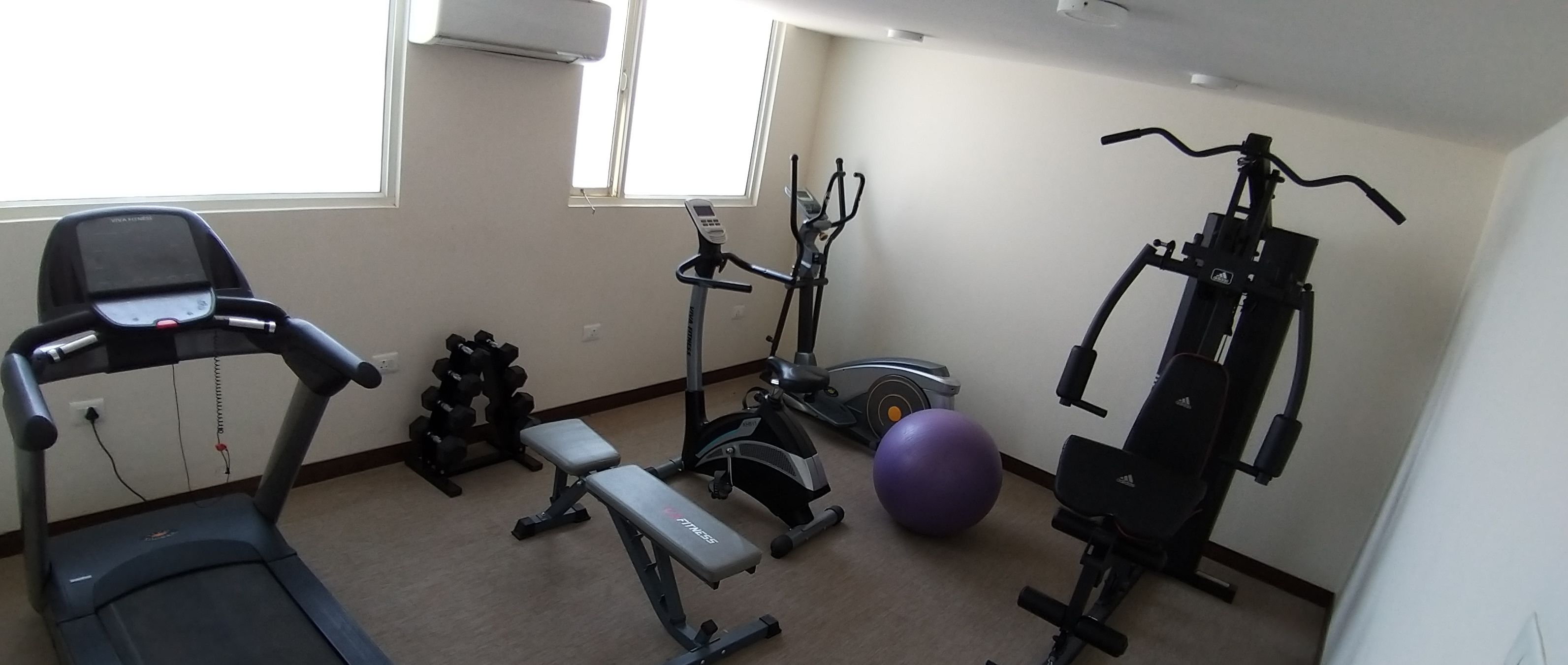 Un gimnasio con cinta de correr, elíptica, banco de pesas, mancuernas y balón medicinal.
