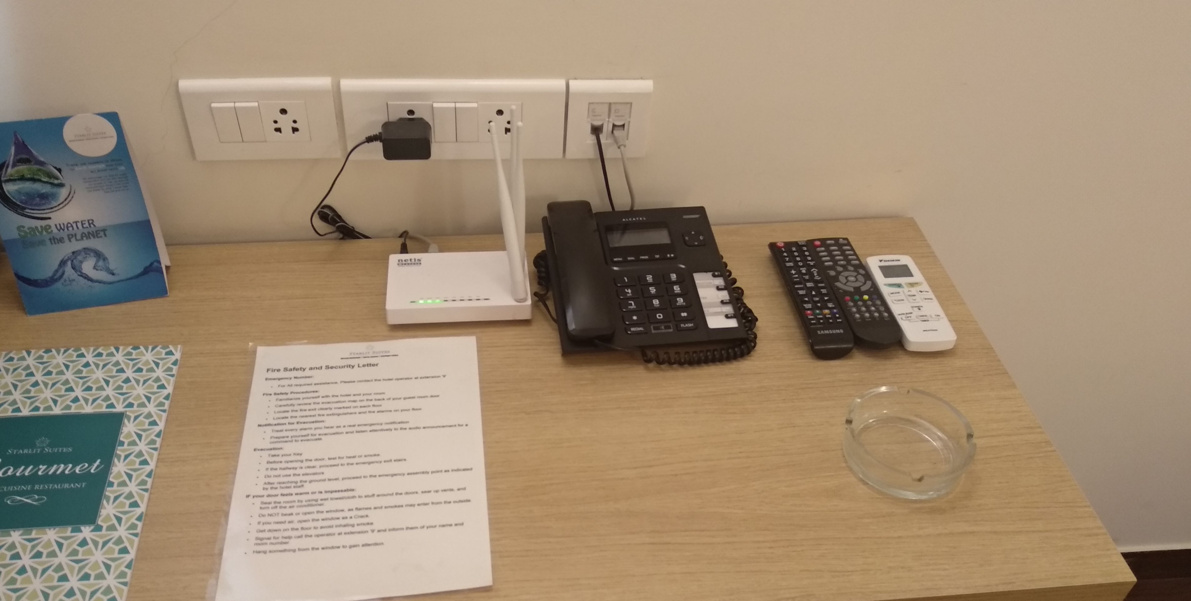 A desk with a Starlet Suites Gourmet restaurant menu, a fire safety and security letter, a Netia router, a Samsung and a Philips phone, and a glass container.