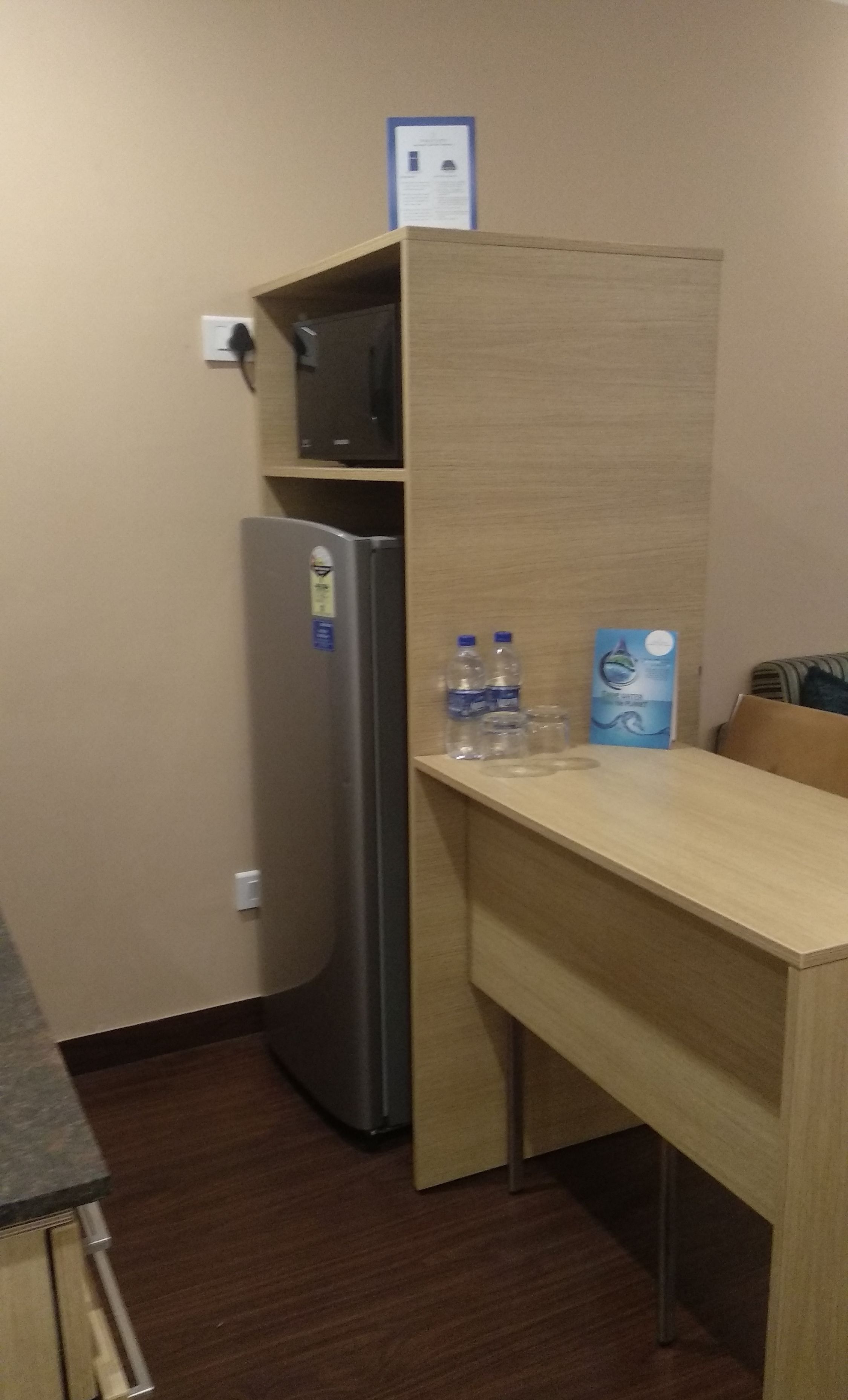 A compact kitchenette with a refrigerator, microwave, and a small shelf. There are two water bottles and a blue informational card on the counter.