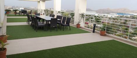 Una terraza en la azotea con comedor, césped artificial, plantas en macetas y vistas de la ciudad.