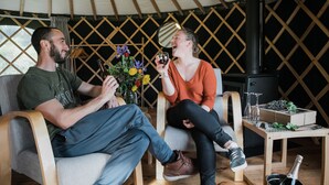Una pareja sentada dentro de una tienda de campaña con paredes de madera, una mesa pequeña con una botella y vasos, y una planta decorativa.