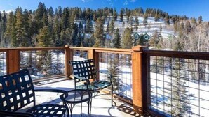 A wooden deck with two chairs and a table, overlooking a snowy landscape with trees and a house in the distance. - Mountain Thunder (Breckenridge)