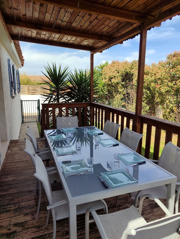 An outdoor patio with a glass-top table and chairs, surrounded by a wooden deck and a wooden pergola.