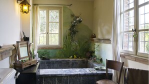A bathroom with a bathtub, a wooden chair, a mirror, and a window with curtains.