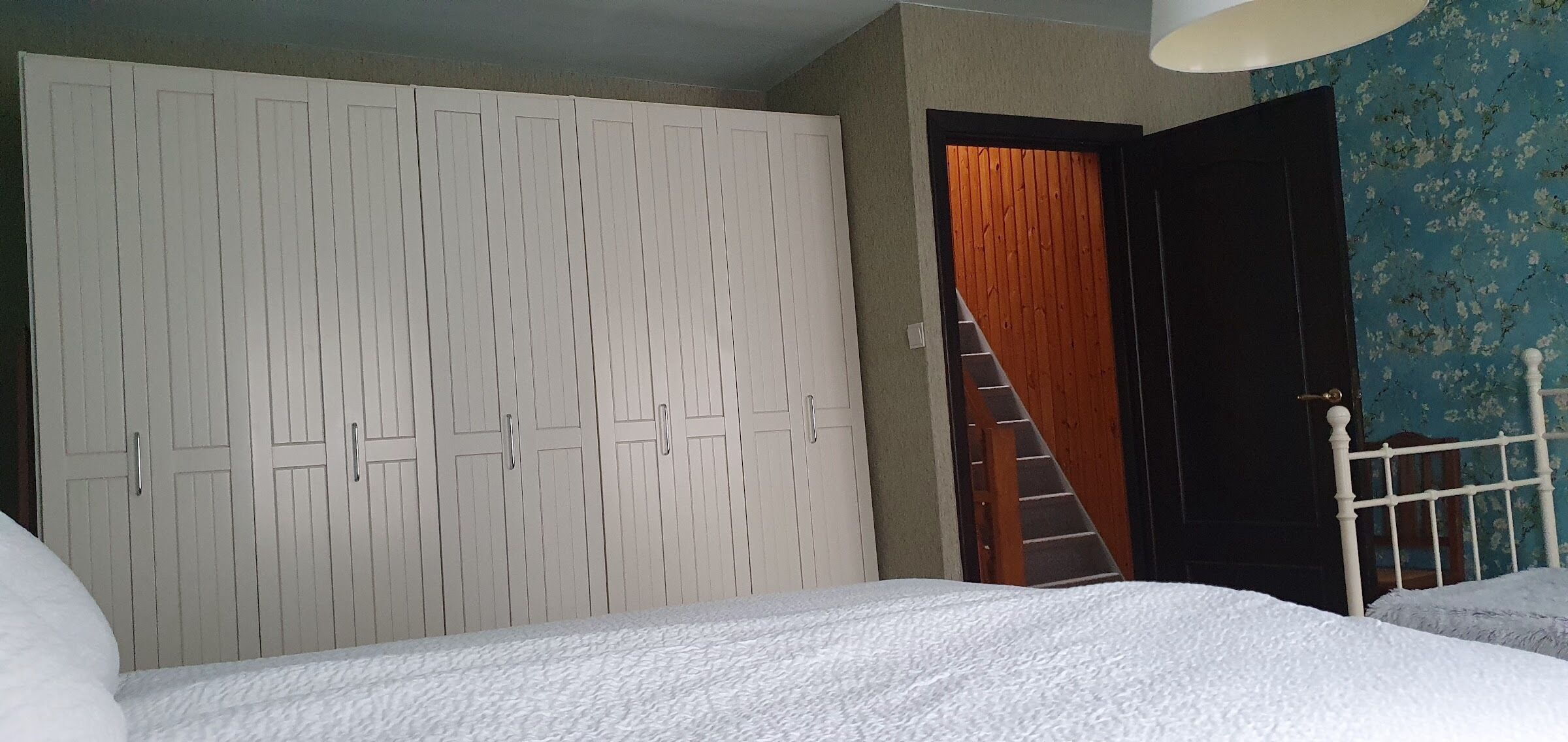 Quarto com cama branca, guarda-roupa branco, porta de madeira e parede com padrão floral azul.