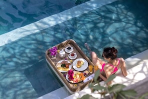 A poolside breakfast setup with a tray of food and drinks, a woman in a pink swimsuit, and a clear view of the pool. - Villa Seriska Jimbaran Bali (Jimbaran)