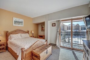 A bedroom with a wooden bed, a nightstand, a dresser, and a large window with a view of buildings.