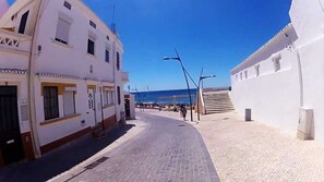 Un carrer estret amb edificis blancs, un cel blau clar i una vista del mar a la distància.