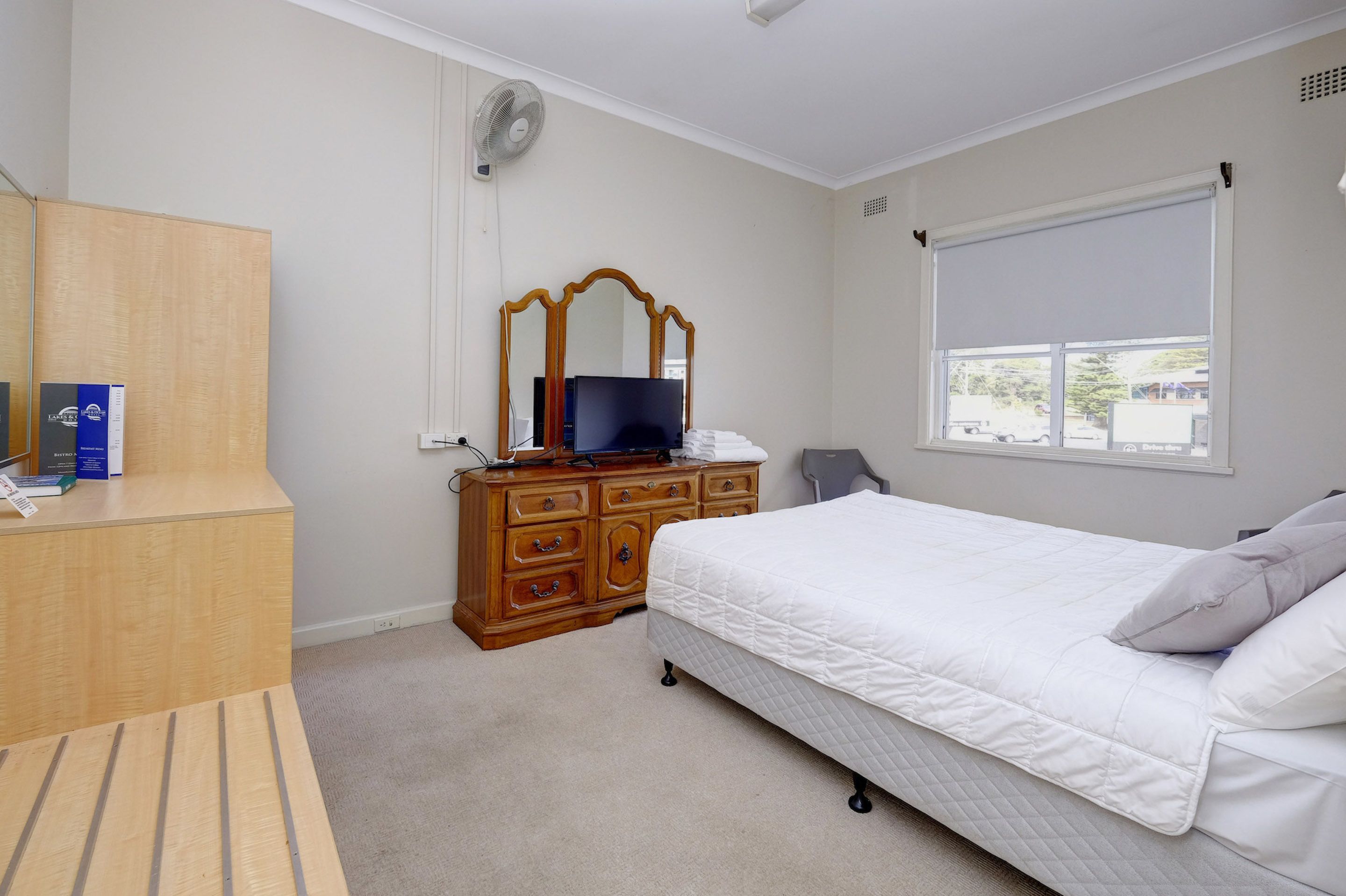 A bedroom with a bed, a wooden dresser, a mirror, a television, and a window with a view of the outside.