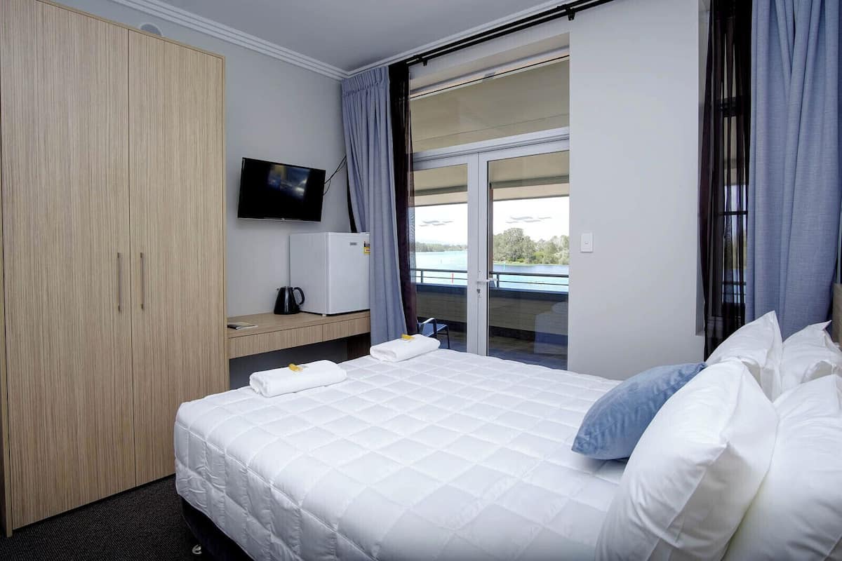 A hotel room with a bed, a television, a small refrigerator, and a view of a body of water through a sliding door.