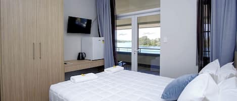 A hotel room with a bed, a television, a small refrigerator, and a view of a body of water through a sliding door.