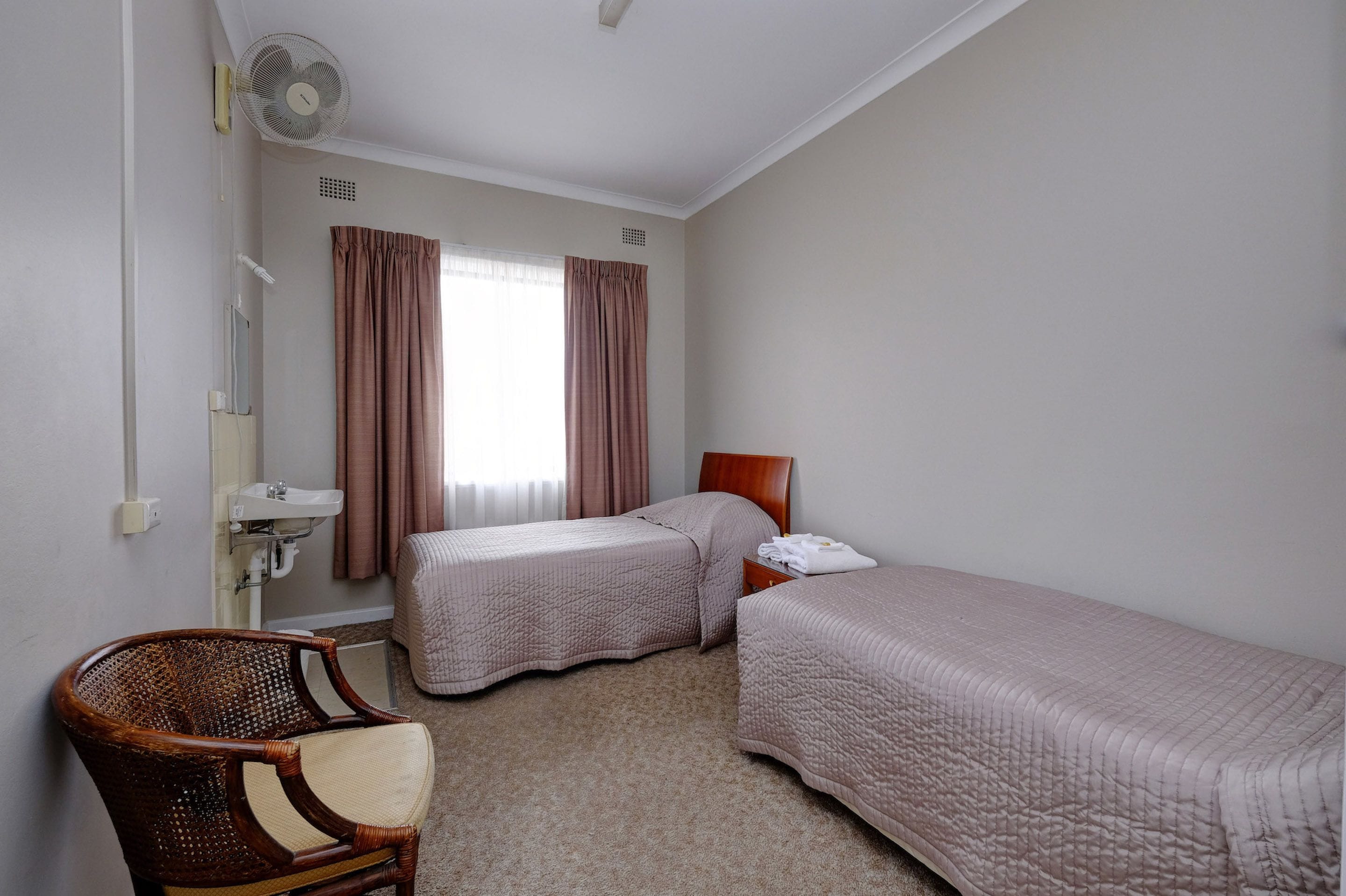 A hotel room with two single beds, a wooden chair, a bedside table, a fan, a window with curtains, and a sink.