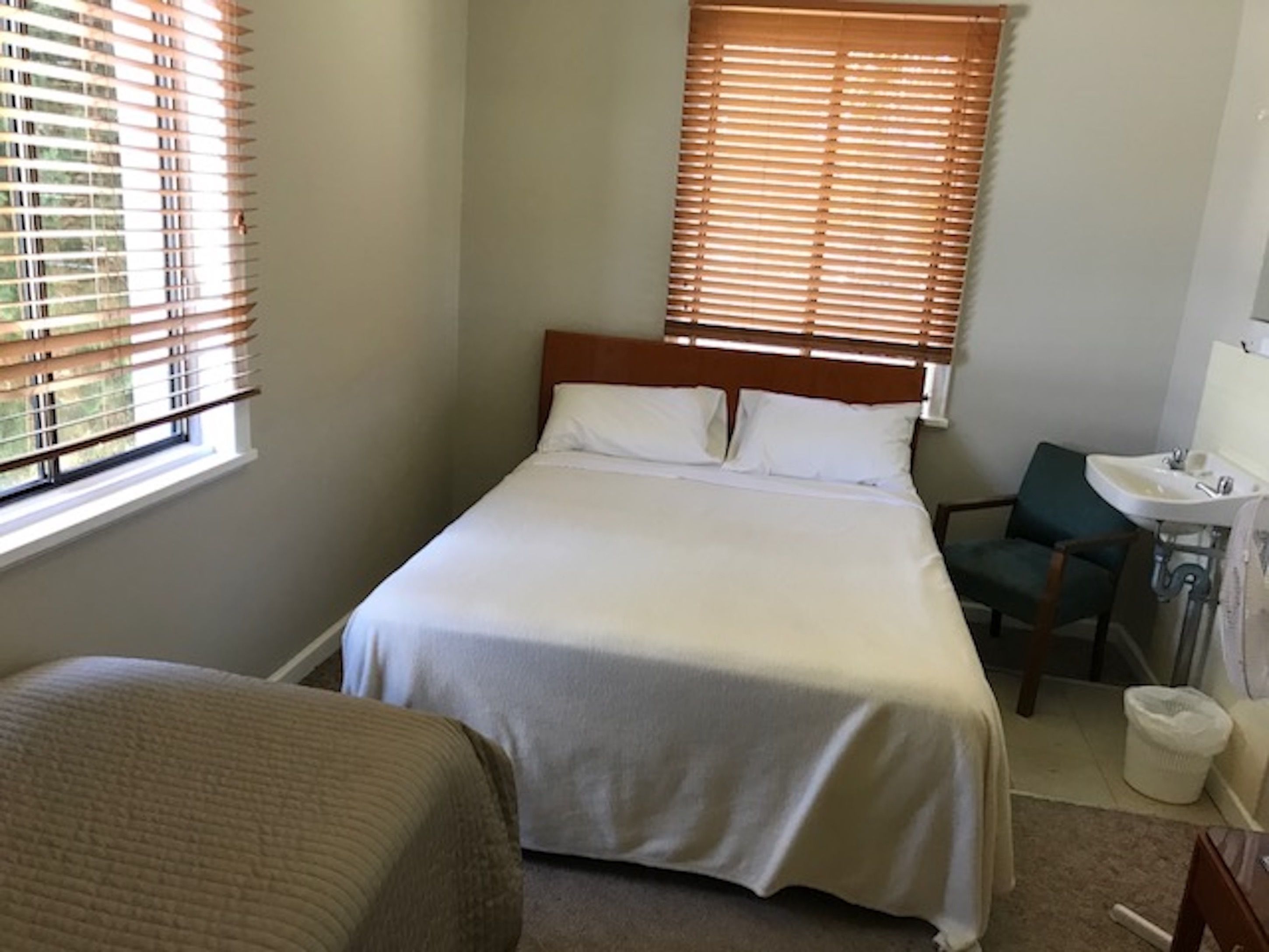 Un dormitorio con cama, una silla, un escritorio pequeño con lavamanos y una ventana con persianas.
