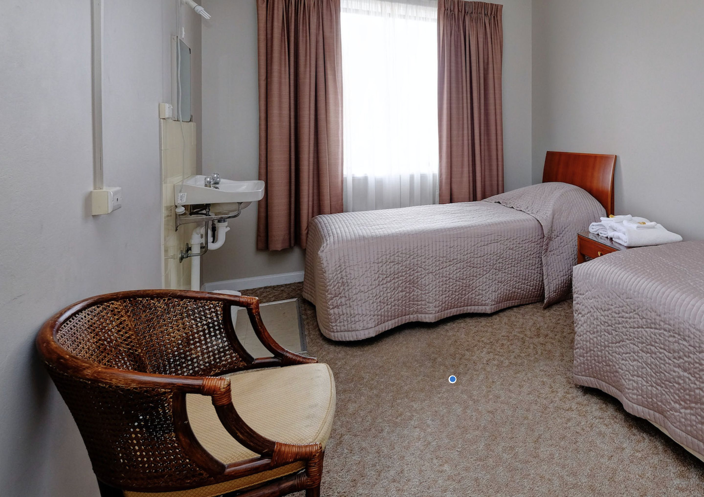 Une chambre d’hôtel avec deux lits, une chaise en bois, un lavabo et une fenêtre avec des rideaux.