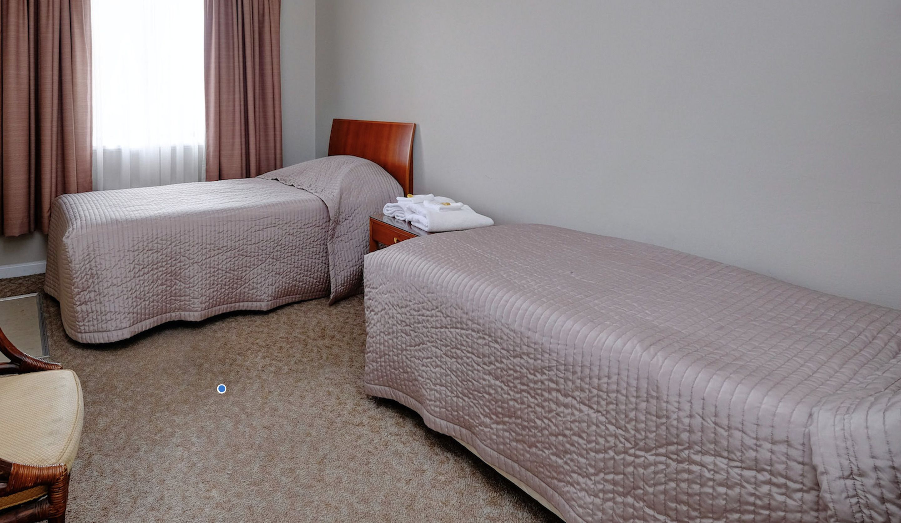 A hotel room with two single beds, a wooden nightstand, a chair, and a window with curtains.